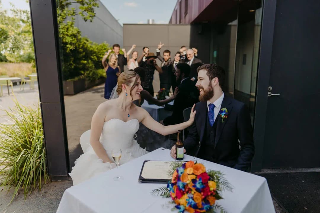 Bride reaches toward the seated groom while they sign wedding paperwork outdoors.