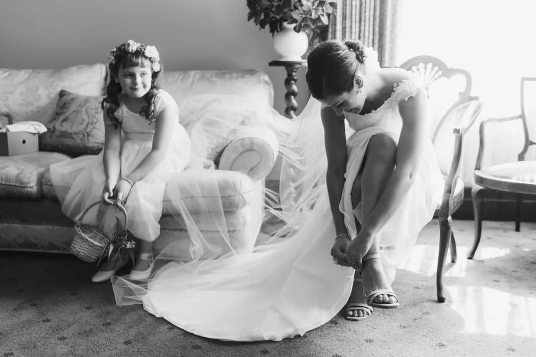 Black and white photograph of the bride fastening her shoe beside a seated flower girl.