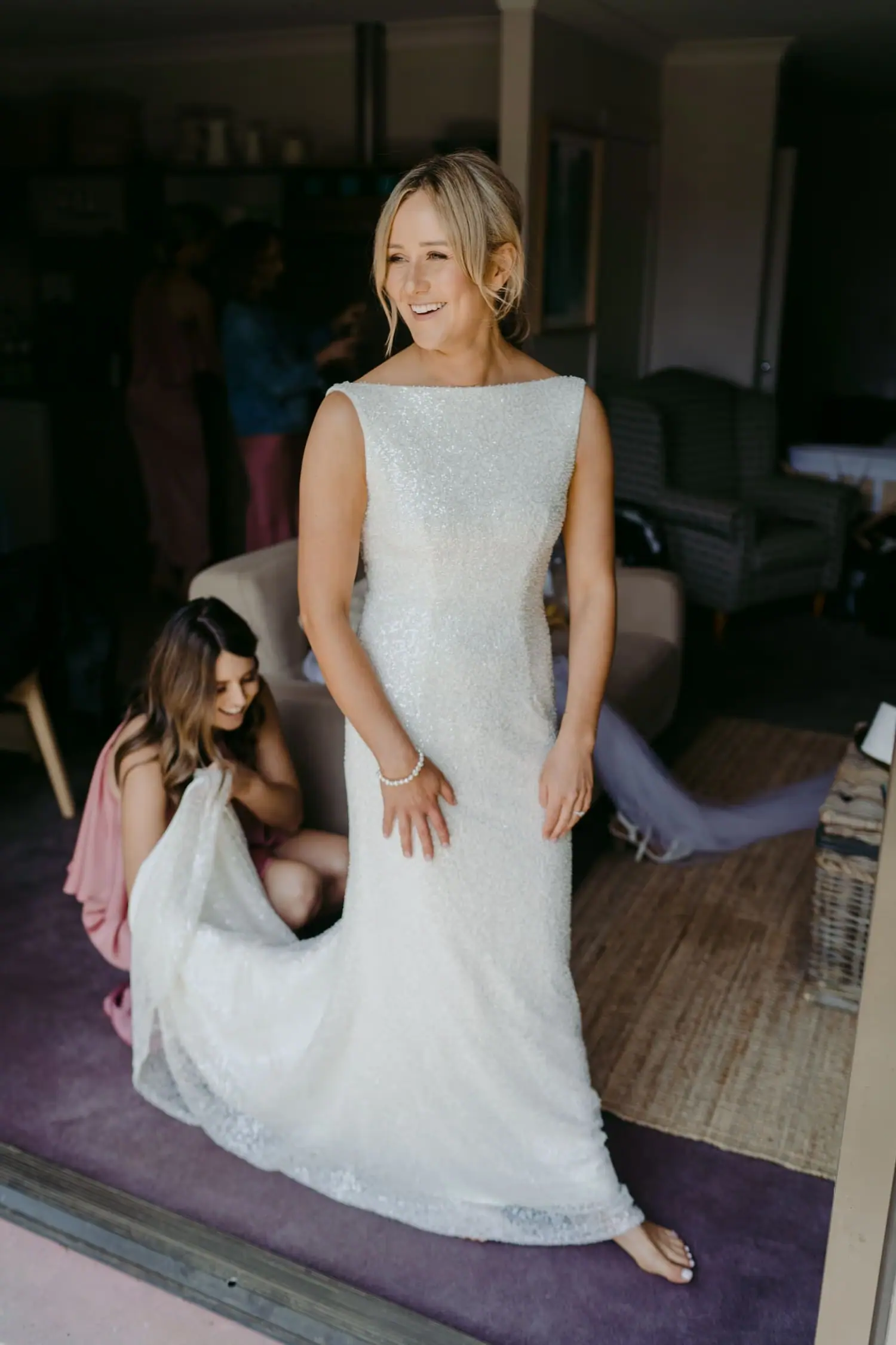 Friend adjusts the bride's dress hem as she stands indoors.