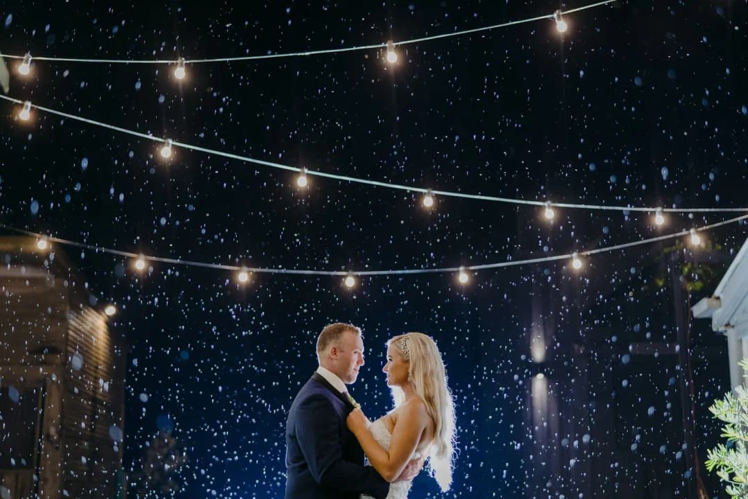 Couple stands together beneath string lights in nighttime rain.
