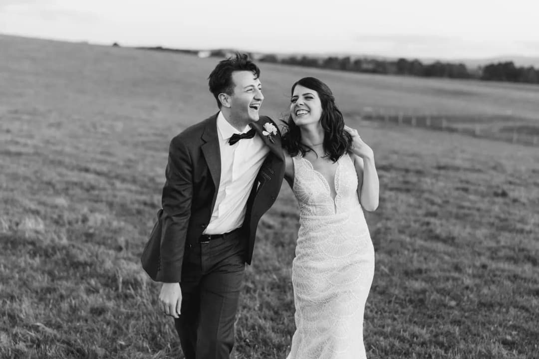 Black and white photograph of the couple laughing while walking through a field.