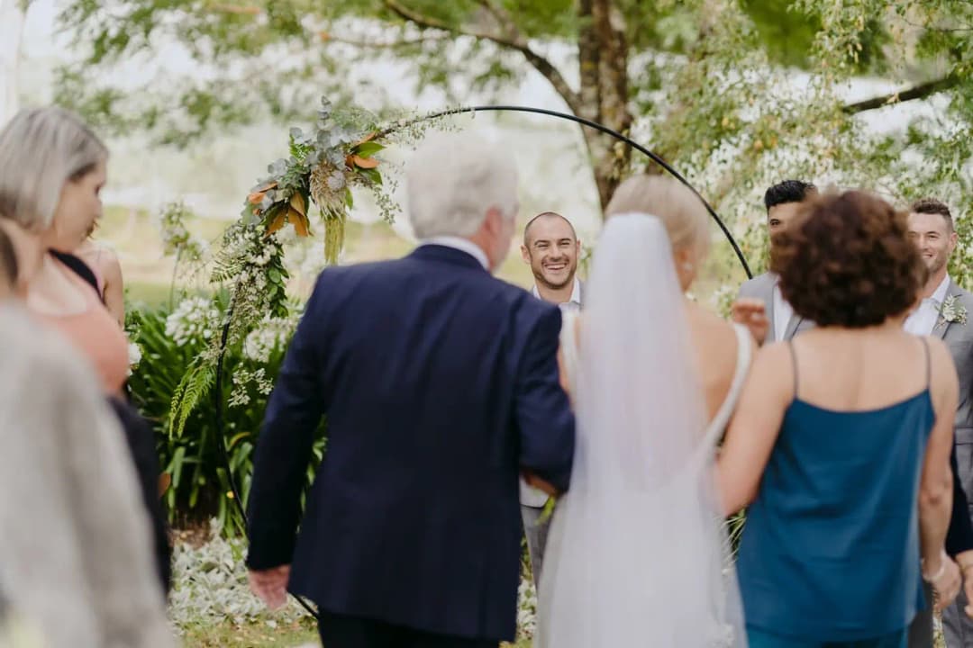 Bride arrives at a circular floral ceremony arch with guests beside her.