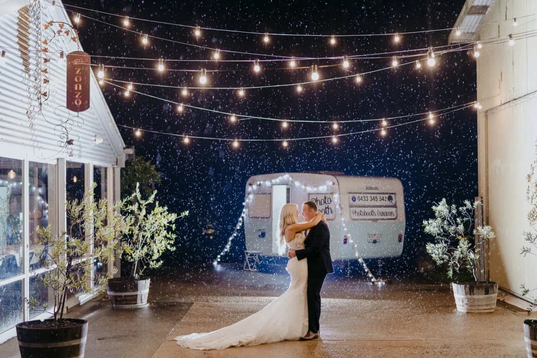 Couple embraces in the rain beneath outdoor string lights with a caravan behind them.