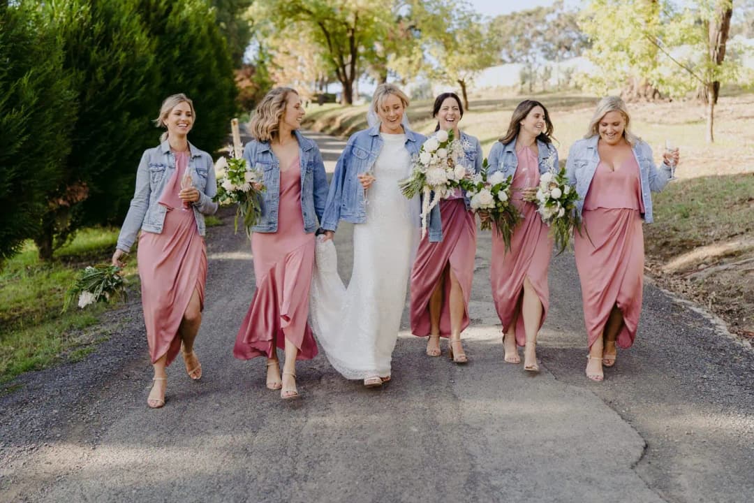 Bride and bridesmaids walk together holding bouquets and wearing denim jackets.