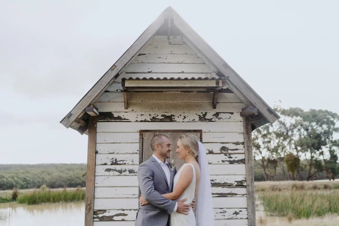 Couple kisses beside a small weathered wooden building overlooking water.