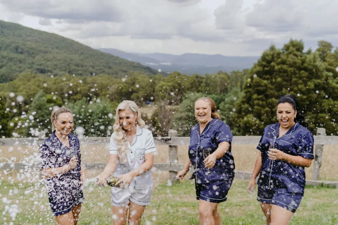 Women in matching pyjamas laugh as champagne sprays outdoors.