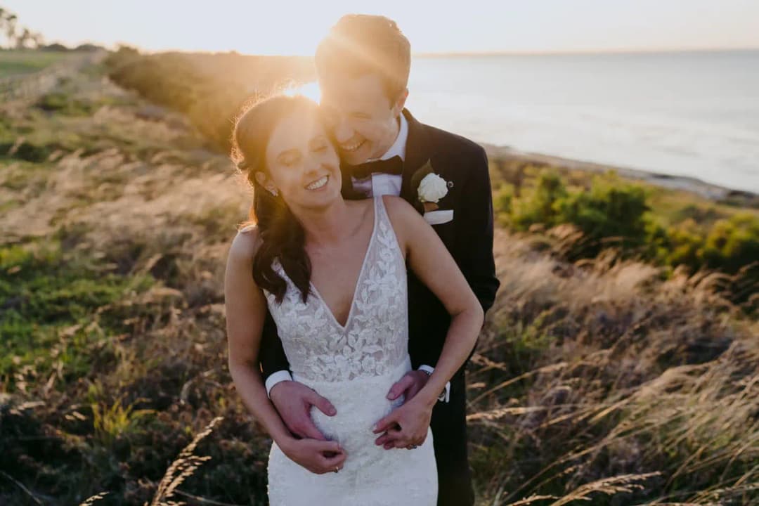 Groom embraces the smiling bride from behind on a grassy coastal hill at sunset.