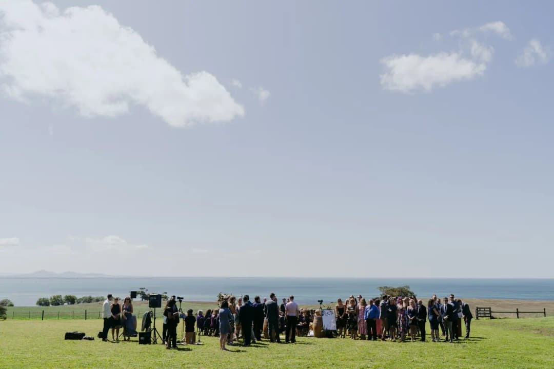 Distant wedding guests gather for a ceremony on a lawn overlooking the sea.