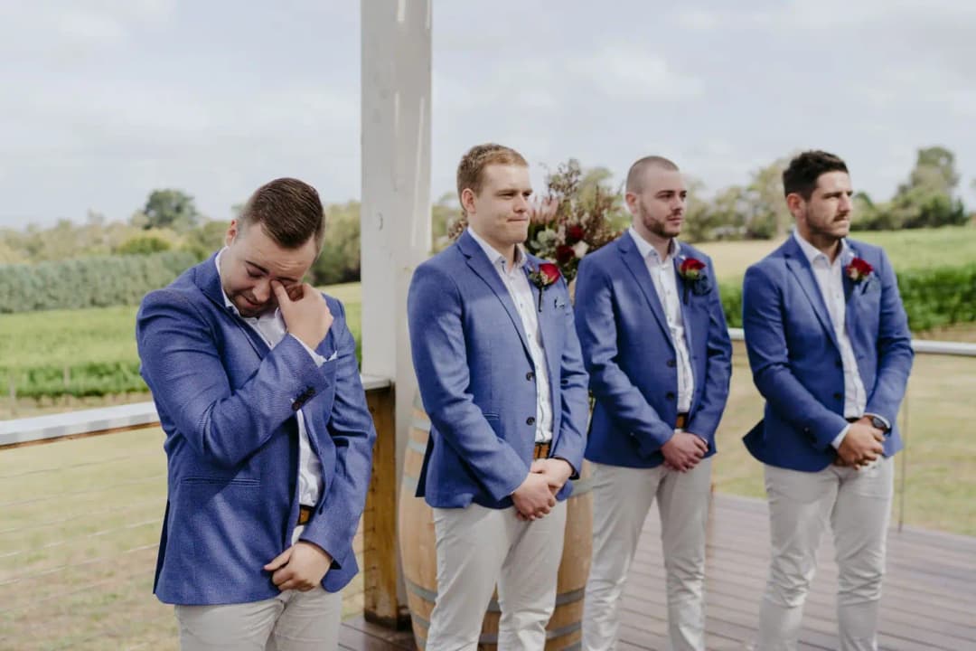 Groom wipes his eye beside three groomsmen in blue jackets.