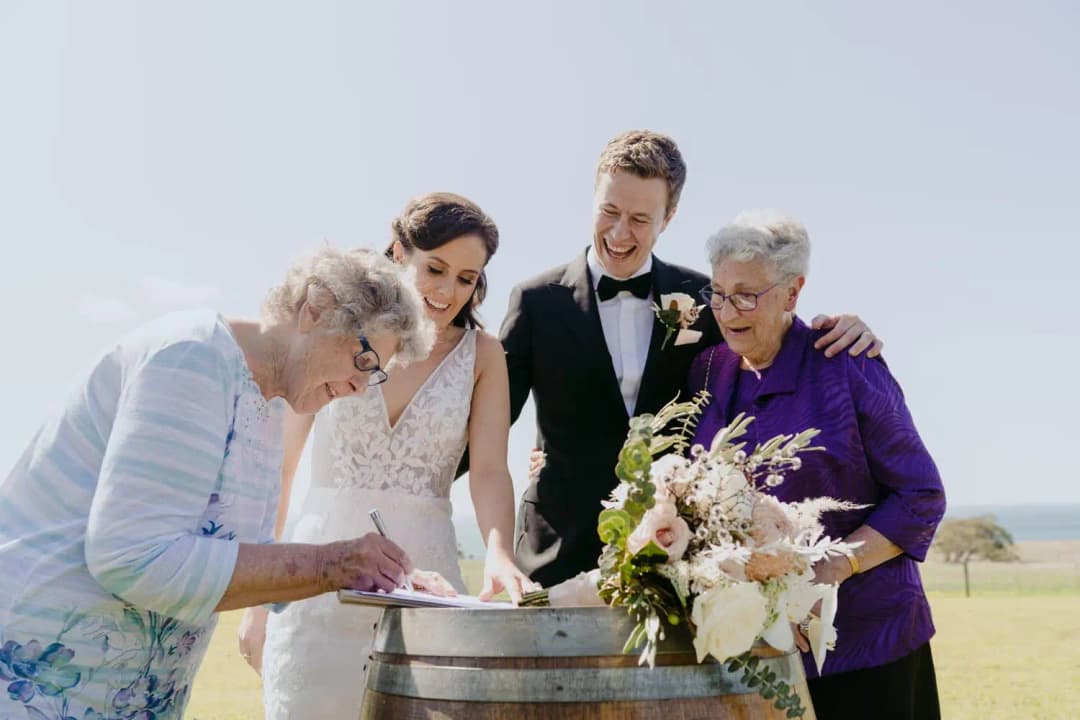 Older woman signs wedding paperwork beside the smiling couple and another guest.