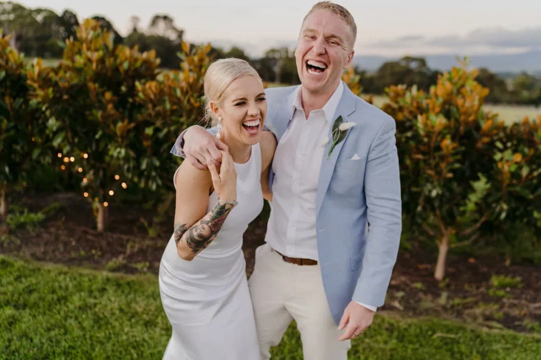 Couple laughs together with their arms around each other beside vineyard foliage.