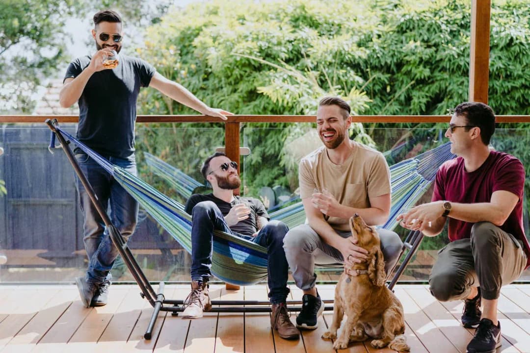 Men relax on a deck beside a hammock with a dog sitting in front of them.