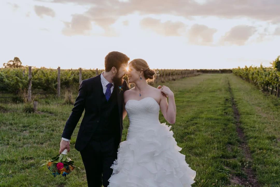 Couple kisses while walking between vineyard rows in warm evening light.