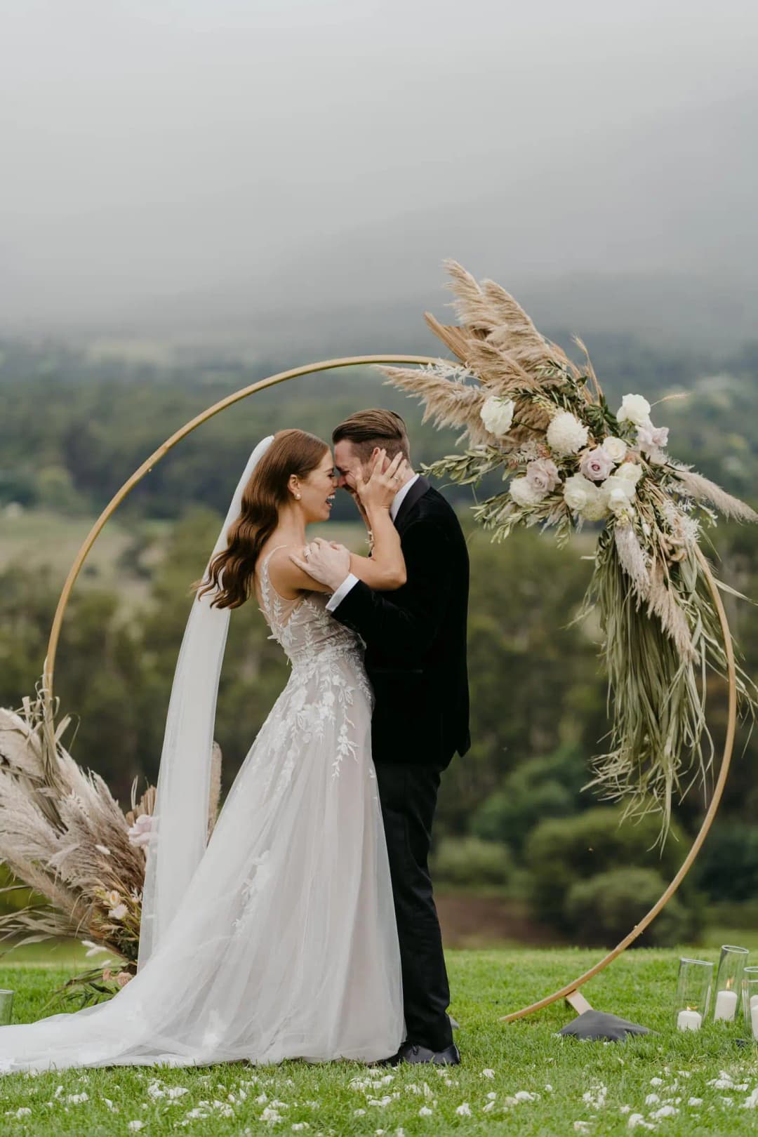 Couple kisses beneath a circular floral arch with misty hills behind them.