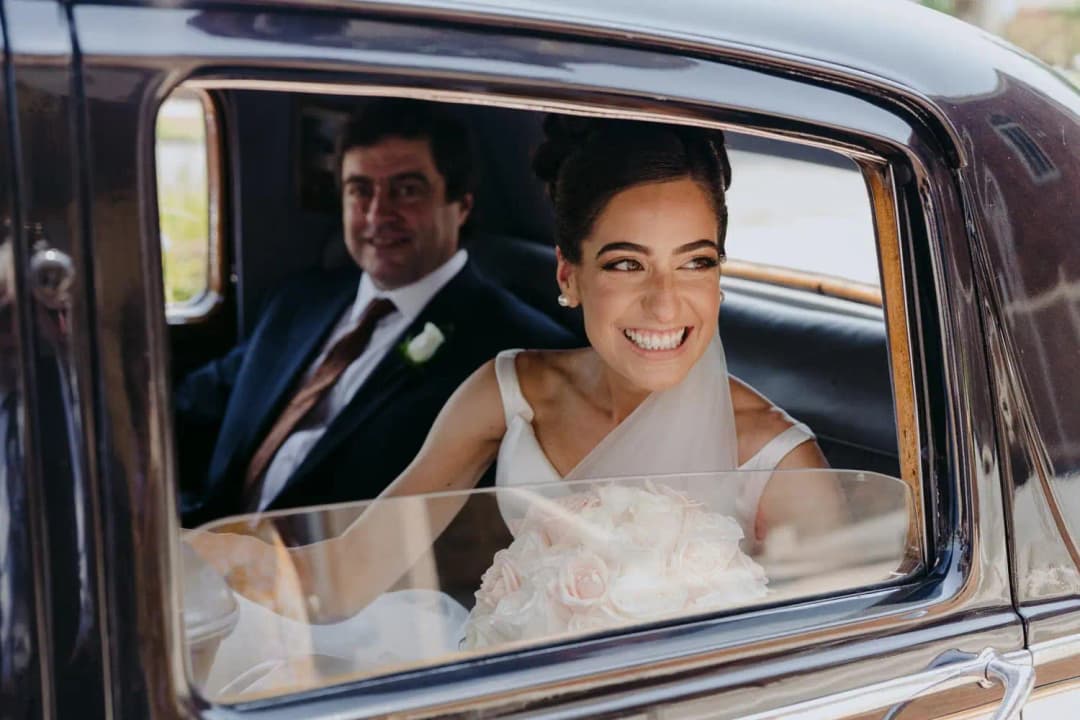 Bride smiles from the window of a vintage car with the groom beside her.