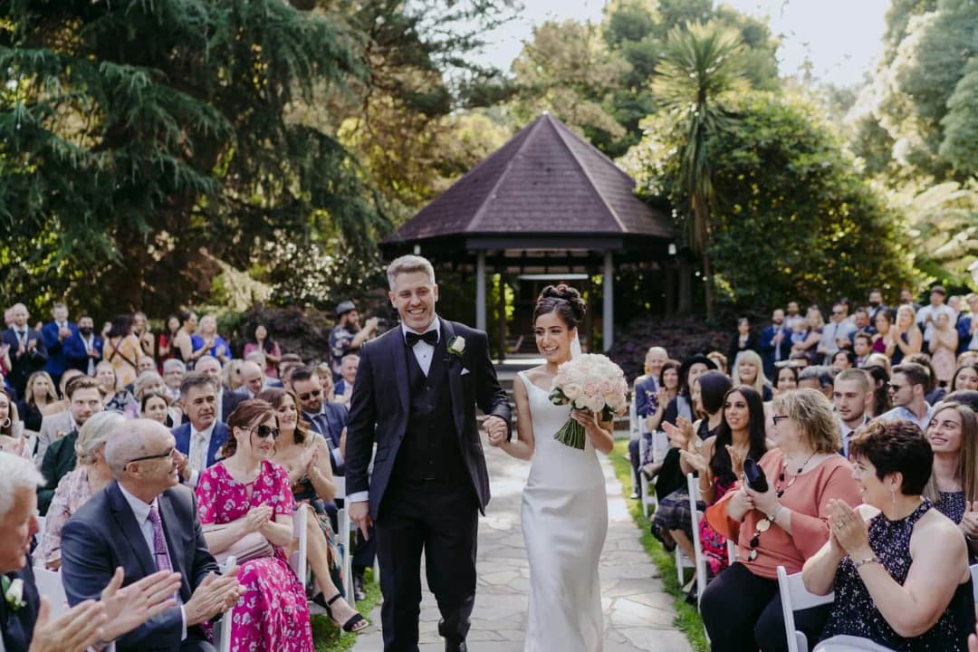 Couple walks down a garden aisle as seated guests applaud.