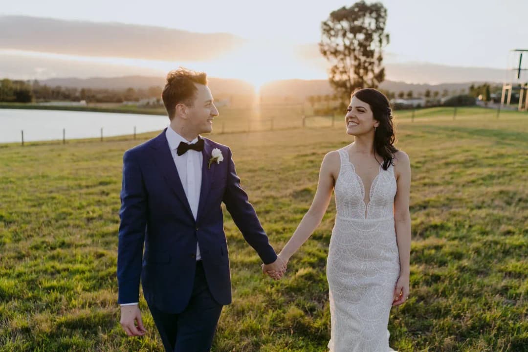 Couple holds hands in a grassy field as the sun sets behind them.