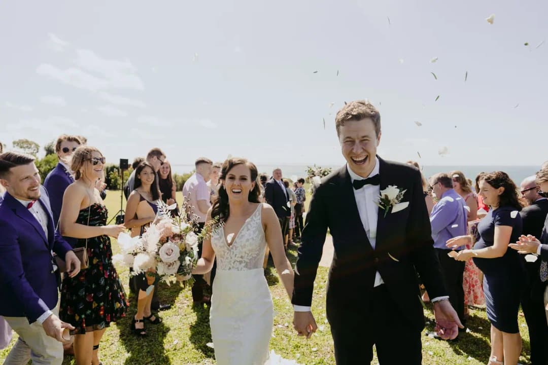 Newlyweds walk hand in hand through applauding guests on a lawn.