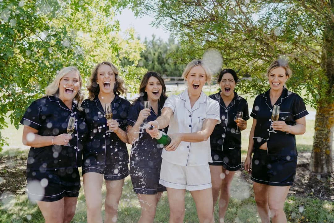 Women in dark and white pyjamas laugh as champagne sprays toward the camera.