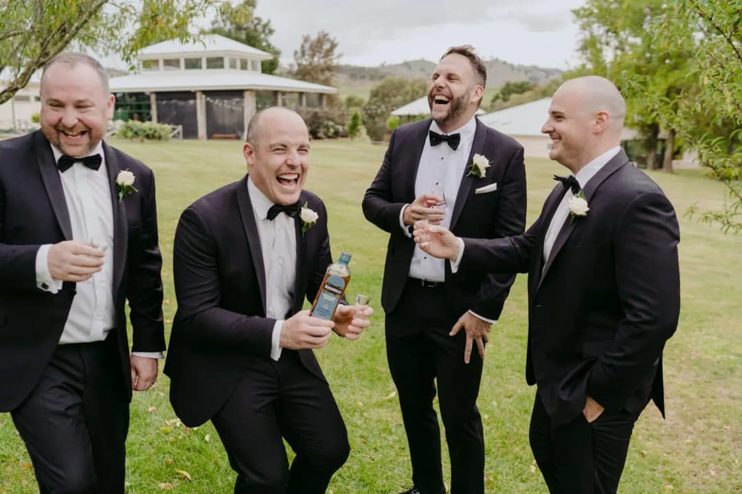 Groom and friends laugh together while holding drinks in a garden.