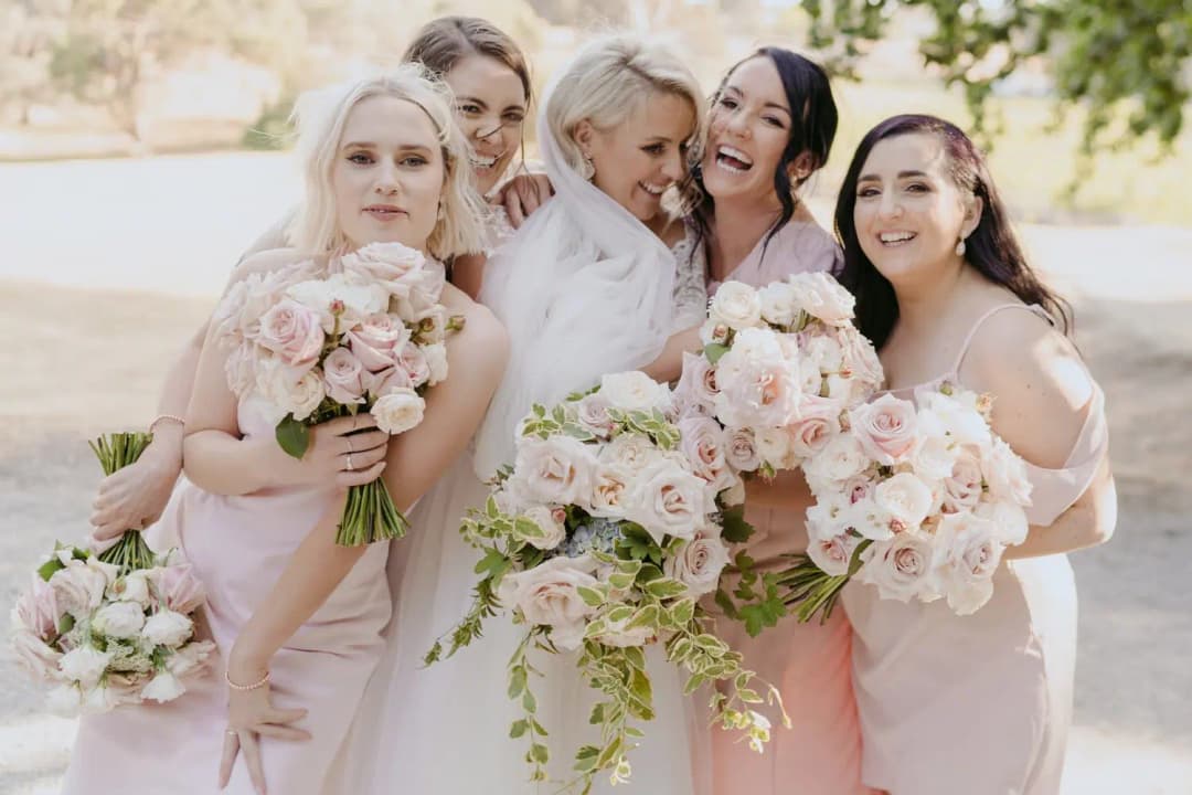Bride and bridesmaids lean together smiling with pink and white bouquets.