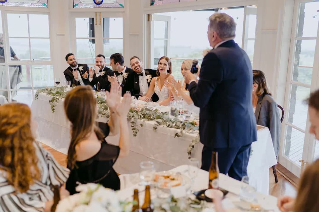 Man gives a speech while the couple and guests applaud at reception tables.