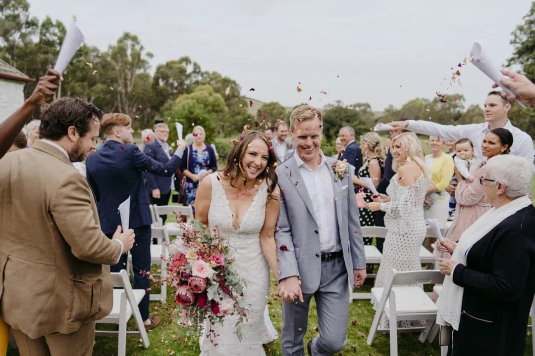 Newlyweds walk through guests throwing confetti beside white ceremony chairs.