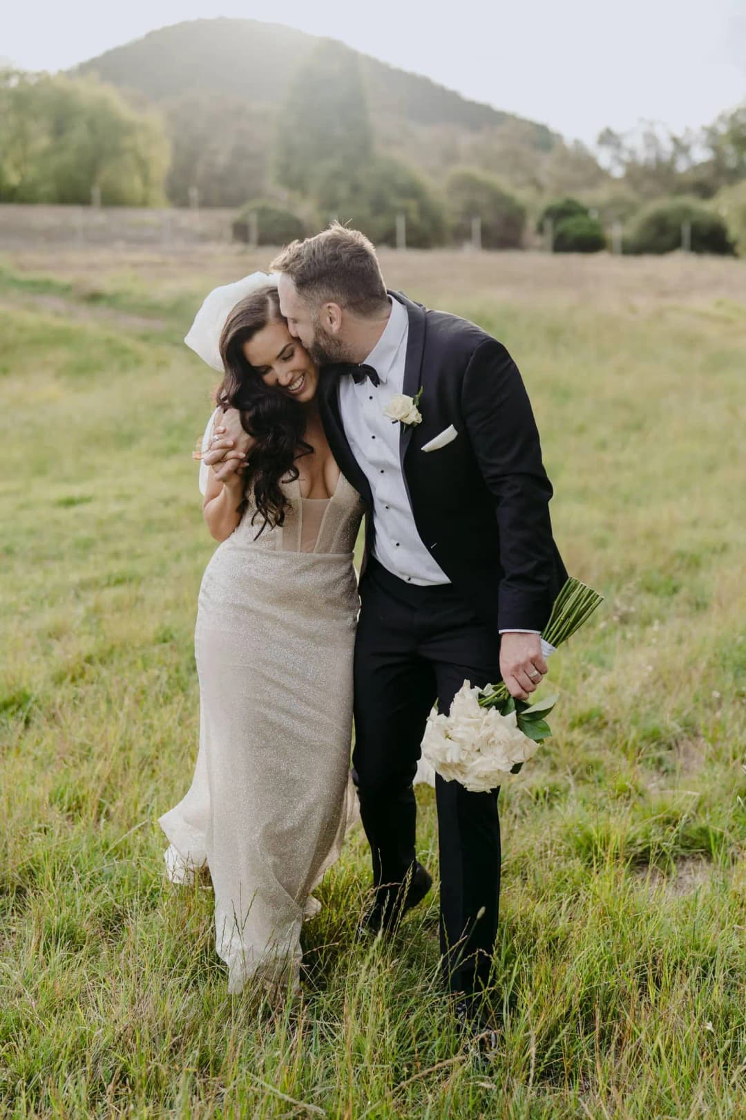 Groom kisses the bride's forehead as they stand together in long grass.