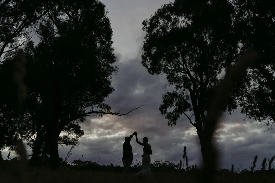Silhouetted couple dances between large trees beneath a dramatic evening sky.