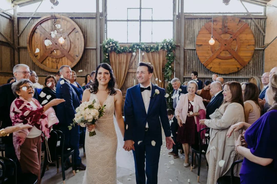 Newlyweds walk through applauding guests in a reception hall with large wooden reels on the wall.