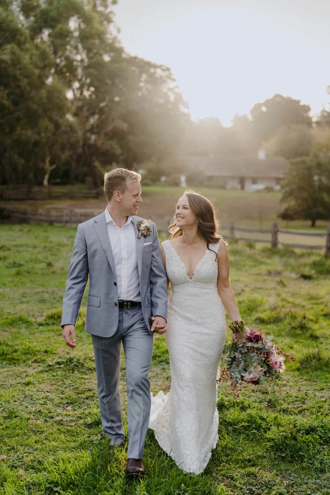 Couple walks hand in hand across sunlit grass holding a bouquet.