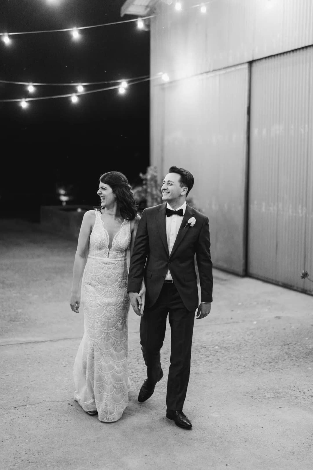 Black and white photograph of the couple walking beneath outdoor string lights at night.