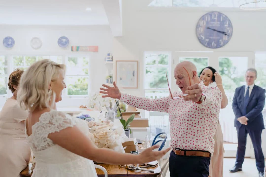 Older man opens his arms to greet the bride indoors.