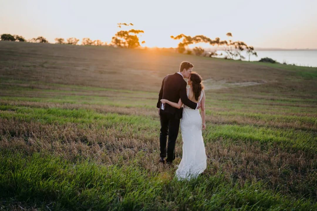 Couple embraces in a field with the setting sun above the distant trees.