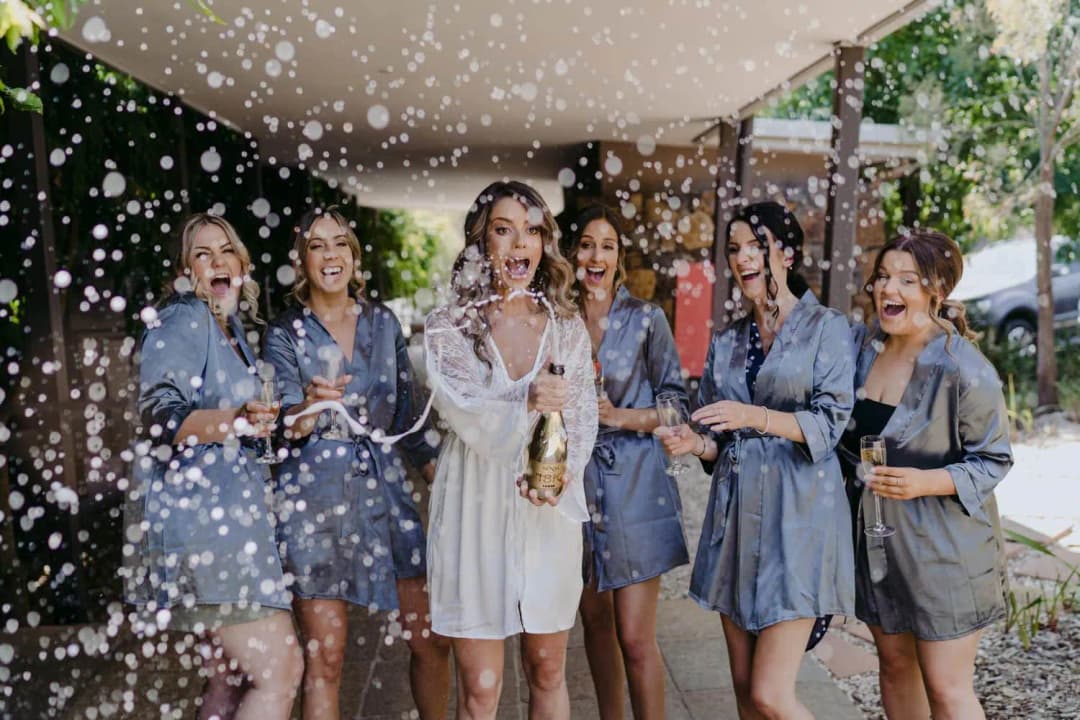Women in white and blue robes laugh as champagne sprays beneath a porch.