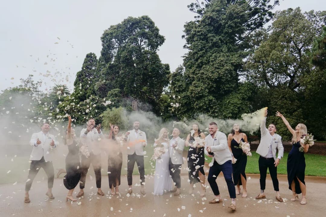 Wedding party celebrates amid airborne confetti and coloured powder outdoors.