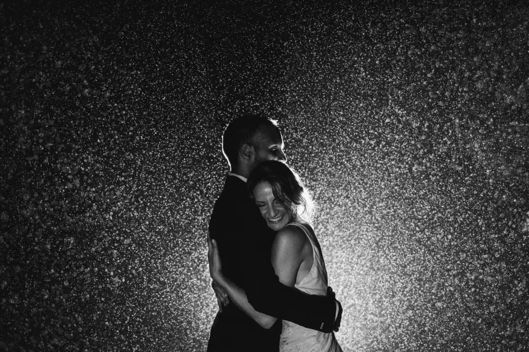 Black and white portrait of the couple embracing in backlit nighttime rain.