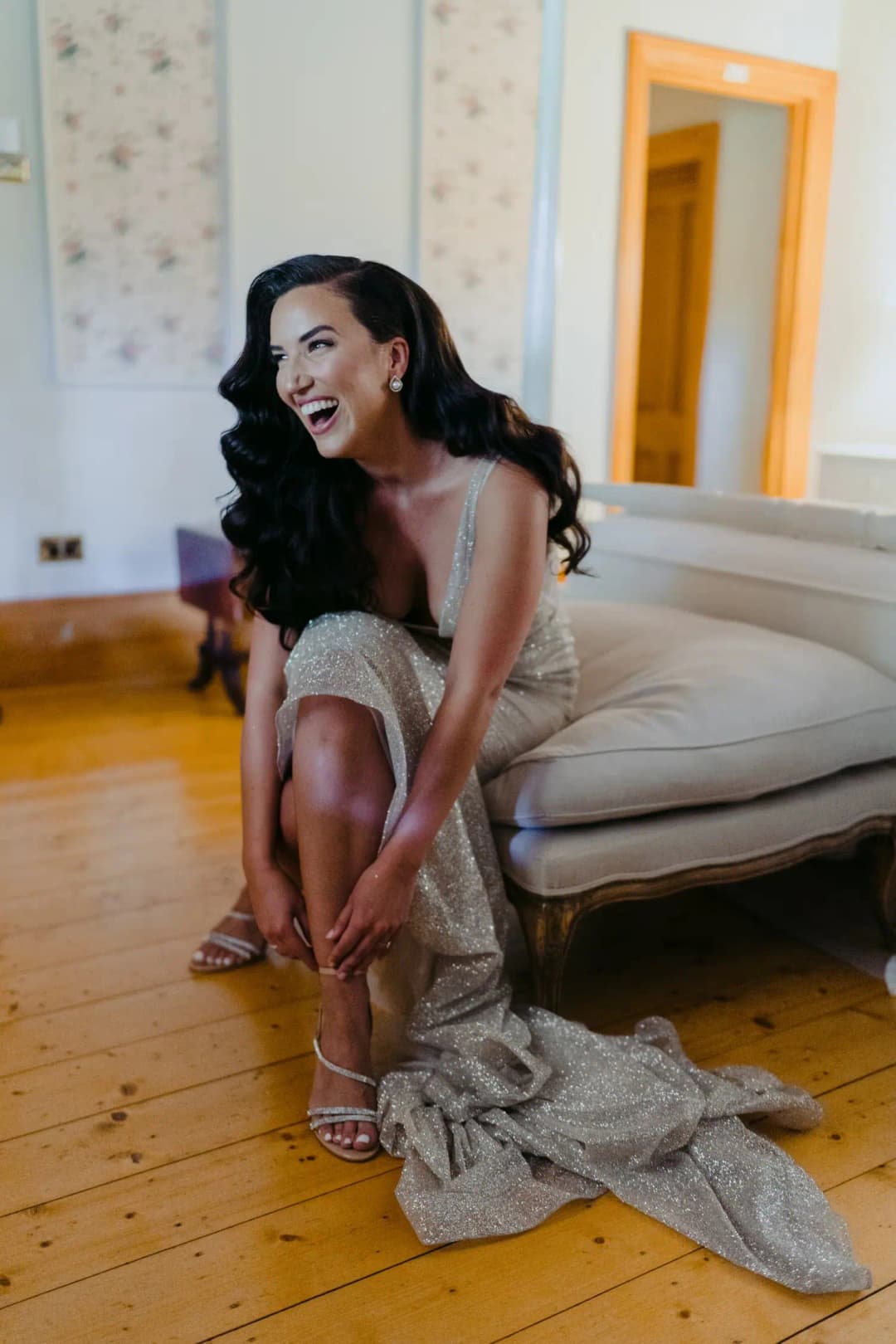 Bride sits laughing as she fastens her shoe beside a sofa.