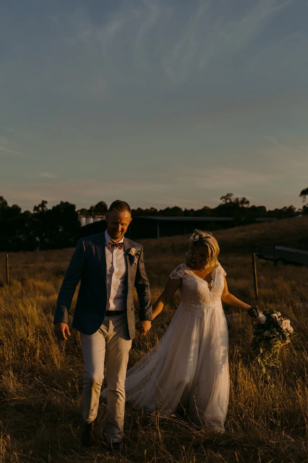 Couple walks through a dry field at dusk with the bride holding her skirt.