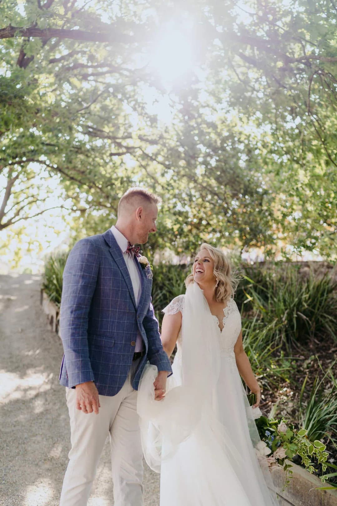 Couple stands beneath sunlit trees as the bride gathers her dress.