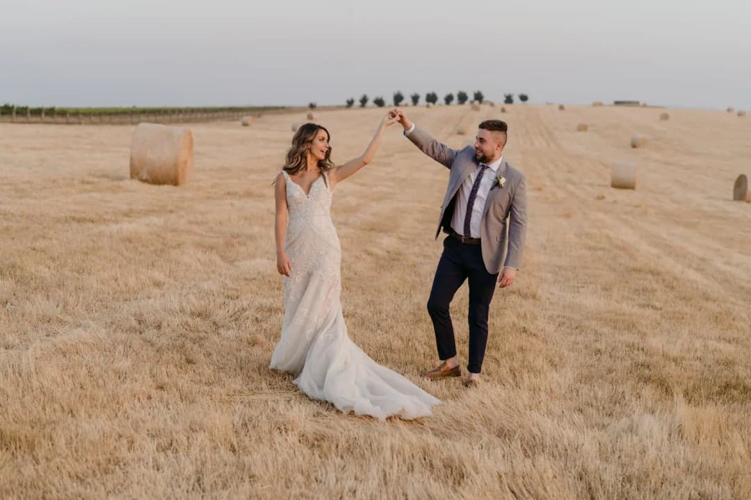 Groom raises the bride's hand for a twirl in a harvested field.