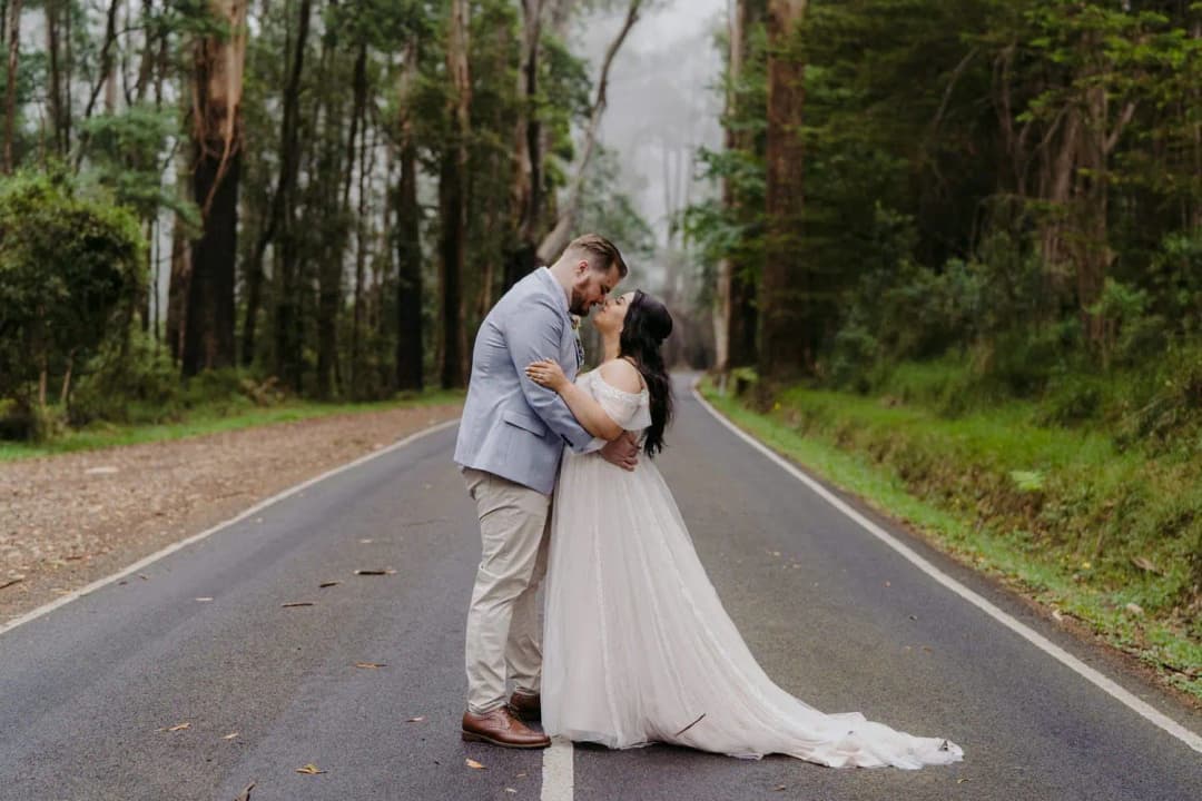 Couple kisses in the middle of a quiet road through tall forest trees.
