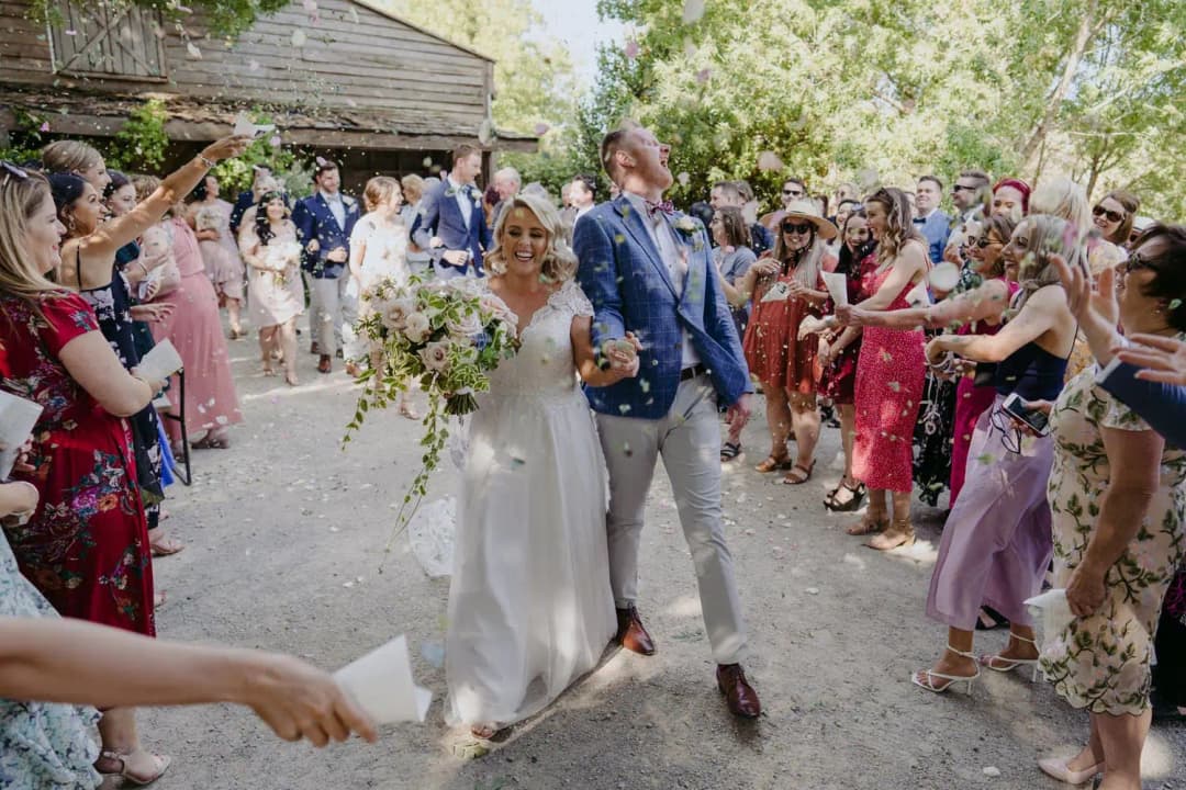 Couple walks through a shower of confetti beside a garden building.