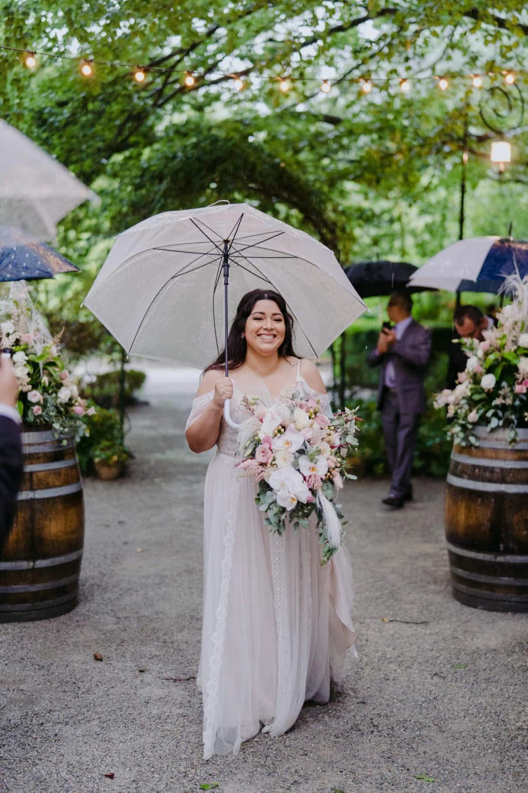 Bride holds a bouquet and clear umbrella on a rainy garden path.