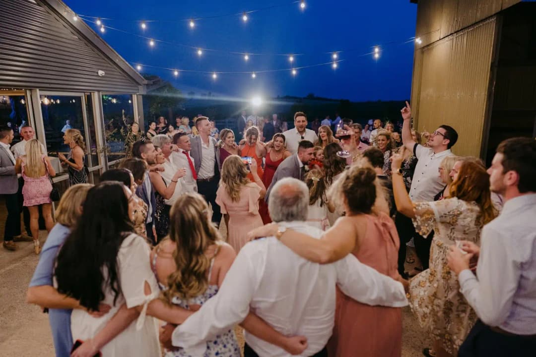 Guests gather closely together beneath nighttime string lights between venue buildings.