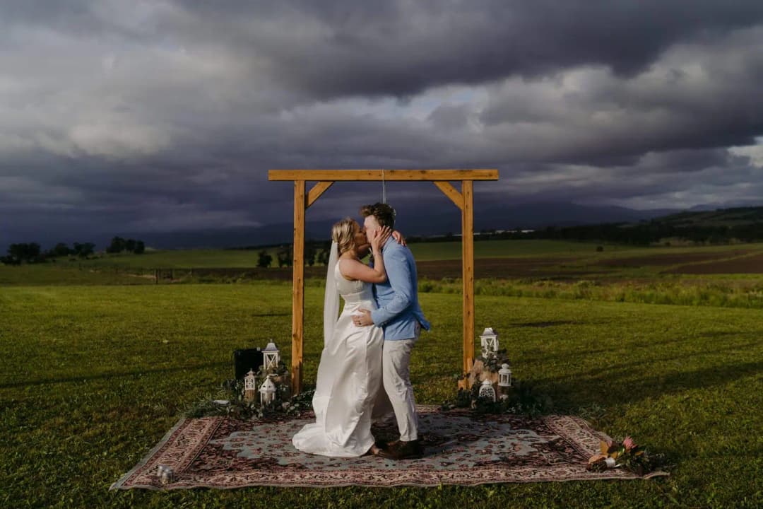 Couple kisses beneath a wooden arch in a field under dark storm clouds.