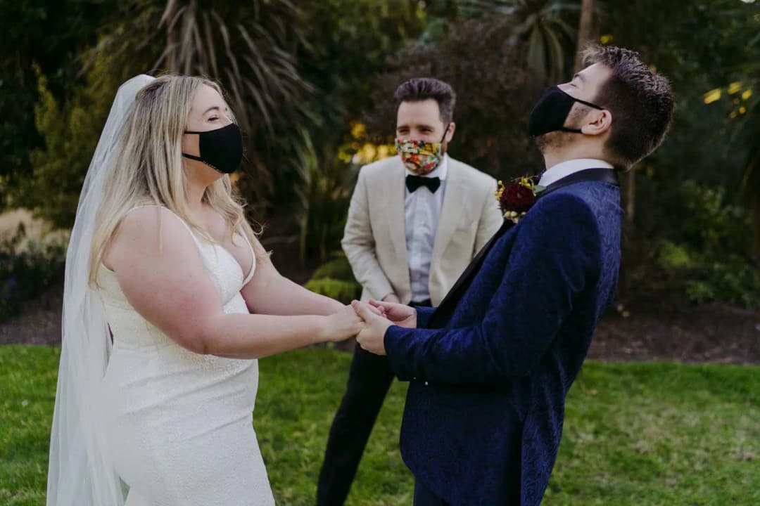 Masked couple holds hands beside a masked celebrant outdoors.