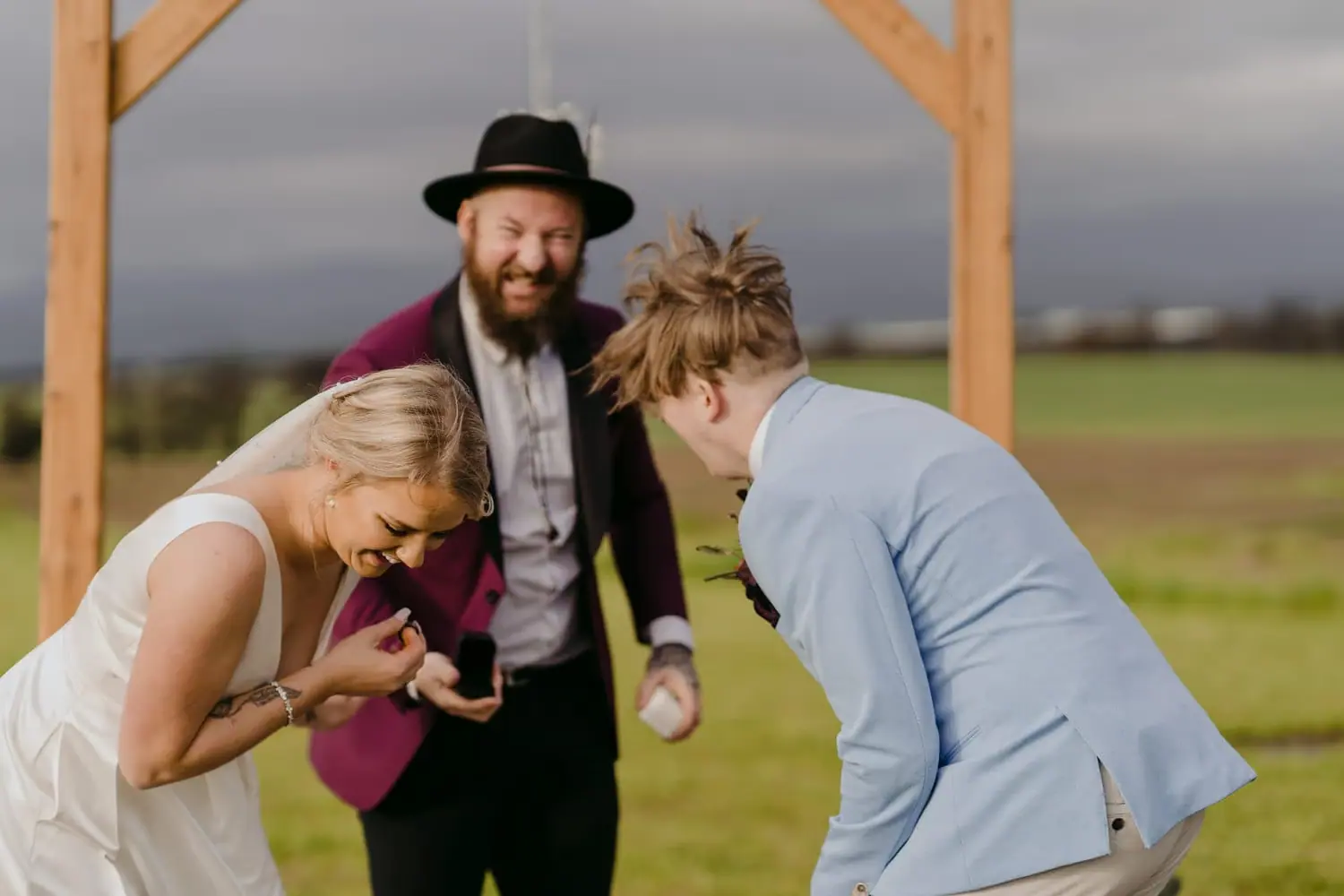 Couple laughs beneath a wooden ceremony arch beside the celebrant.