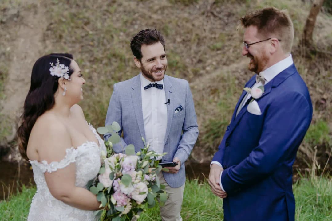Bride and groom smile toward the celebrant beside a wooded creek.