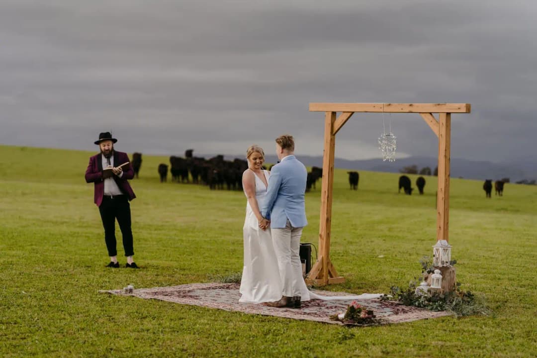 Couple holds hands beneath a wooden arch with cattle in the field behind them.